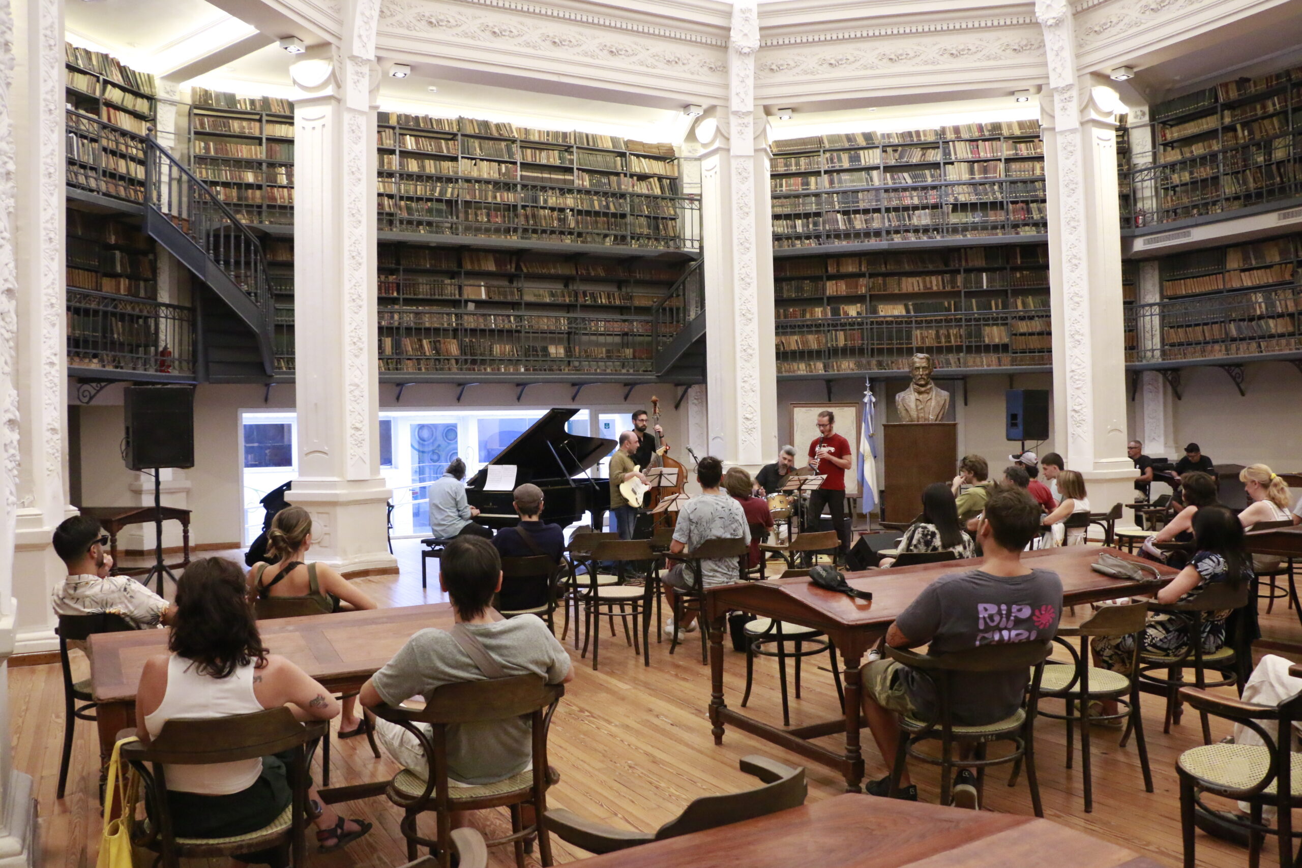 Foto de un grupo de música integrado por cinco personas en concierto en la Sala de lectura Alfredo Lovell donde se encuentran además personas sentadas viendo hacia el grupo.