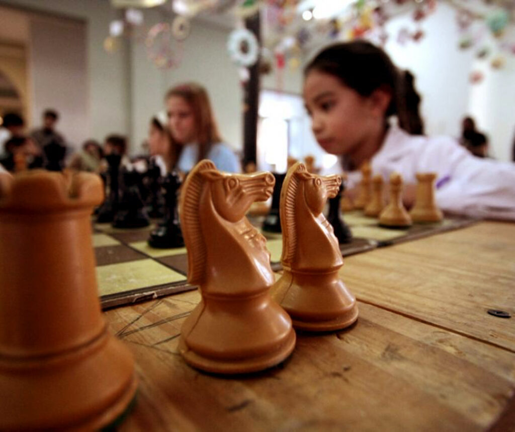 En la foto se observan de una forma difuminada en primer plano piezas de ajedrez, dos caballos, y, fuera de foco, una torre y un tablero con más piezas sobre una mesa de madera y, además, la imagen de una niña jugando el juego y más personas de fondo.
