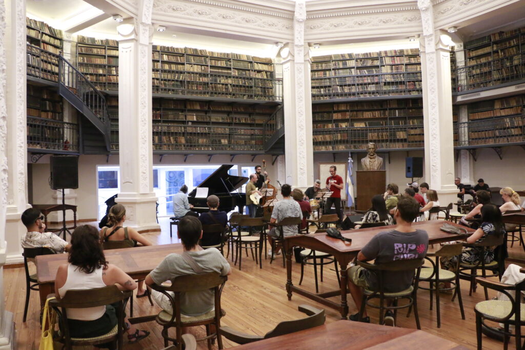 Foto de un grupo de música integrado por cinco personas en concierto en la Sala de lectura Alfredo Lovell donde se encuentran además personas sentadas viendo hacia el grupo.