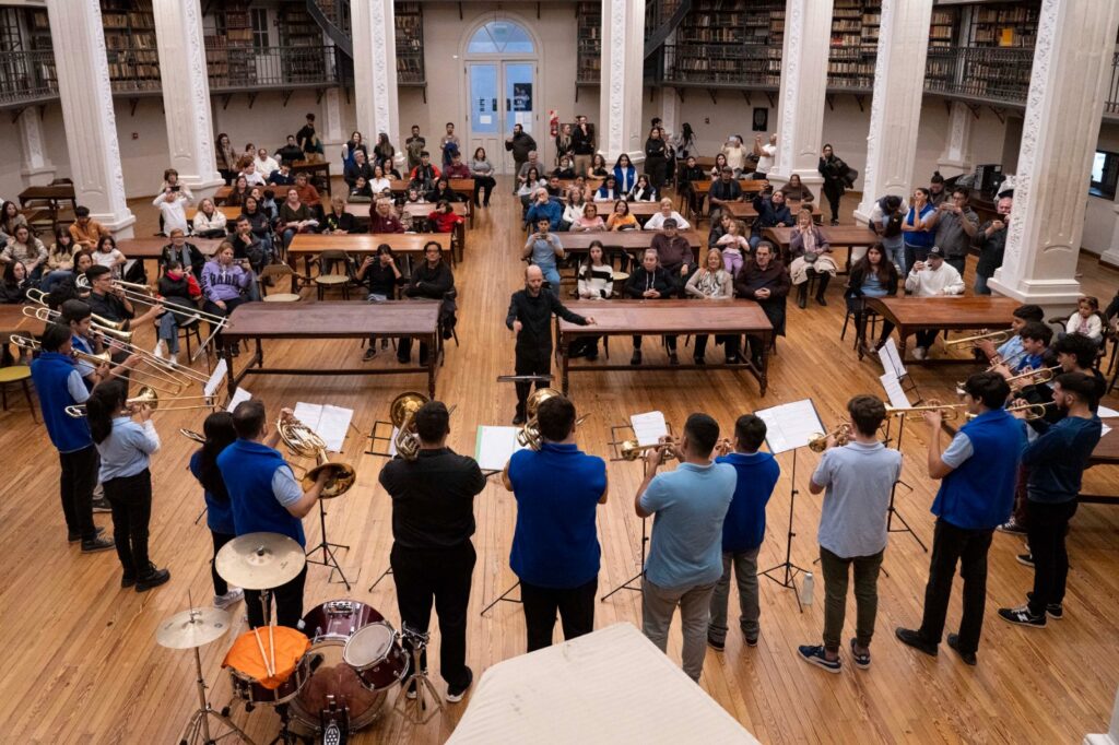 Foto de una orquesta donde se encuentra un conjunto de músicos interpretando una obra musical con diversos instrumentos y bajo la guía de un director con personas sentadas y estanterías de libros de fondo.