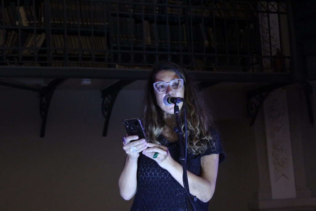 Foto de una mujer iluminada por una luz desde abajo que se encuentra frente a un micrófono con un celular en sus manos, como leyendo, en un ambiente a oscuras con estanterías de libros de fondo.