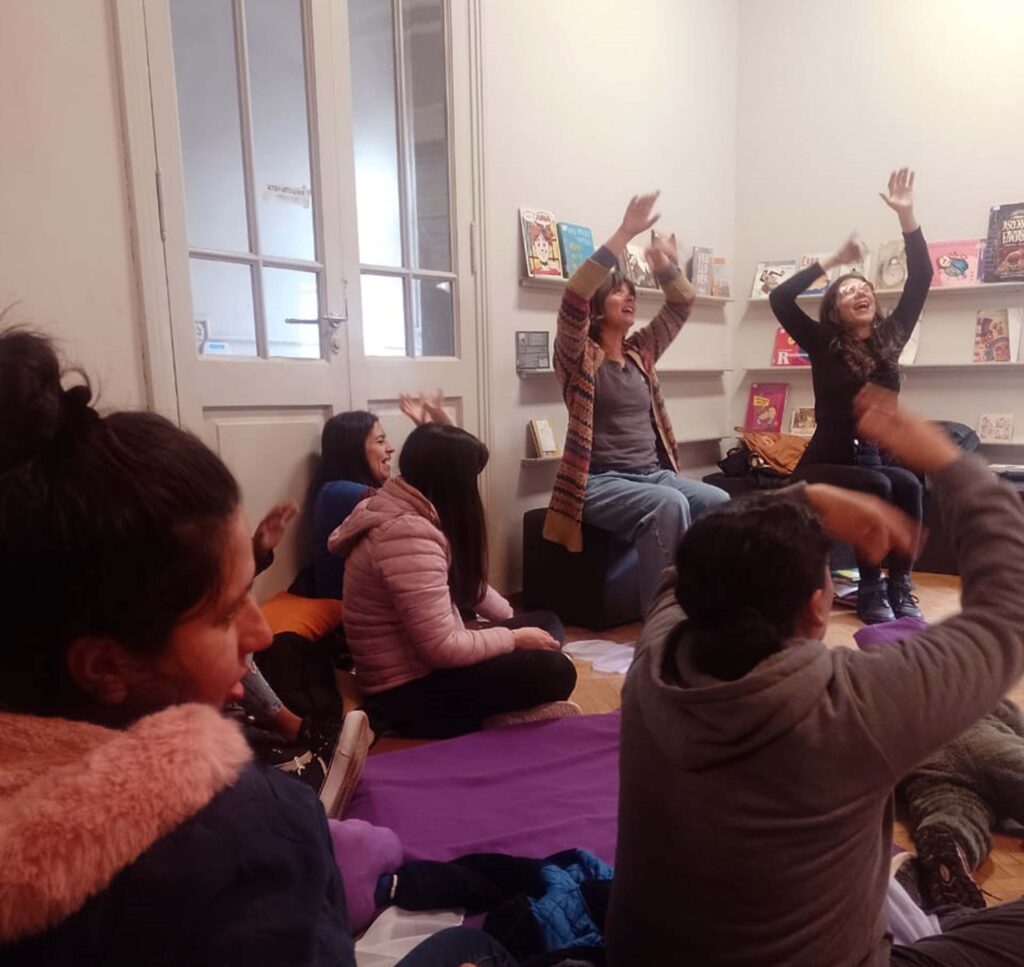 Se observan 2 mujeres sentadas en bancos con sus brazos extendidos hacia arriba. Y, a su alrededor, otras mujeres observándolas y haciendo también gestos con sus brazos hacia arriba.